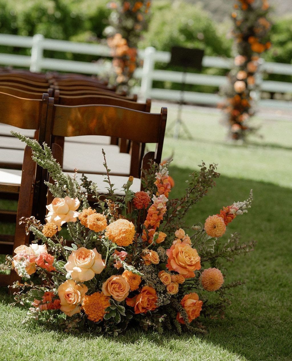 Orange and peach floral arrangement next to wooden chairs on a green lawn.