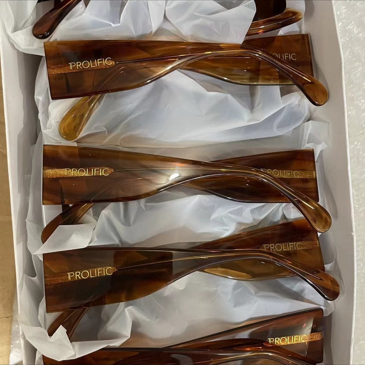 Brown tortoiseshell sunglasses with gold personalized text resting on white tissue paper.