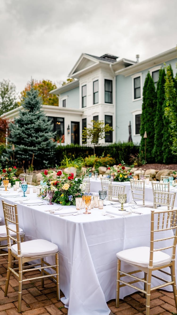 Gold Chiavari chairs with white cushions arranged around long white wedding tables outdoors.