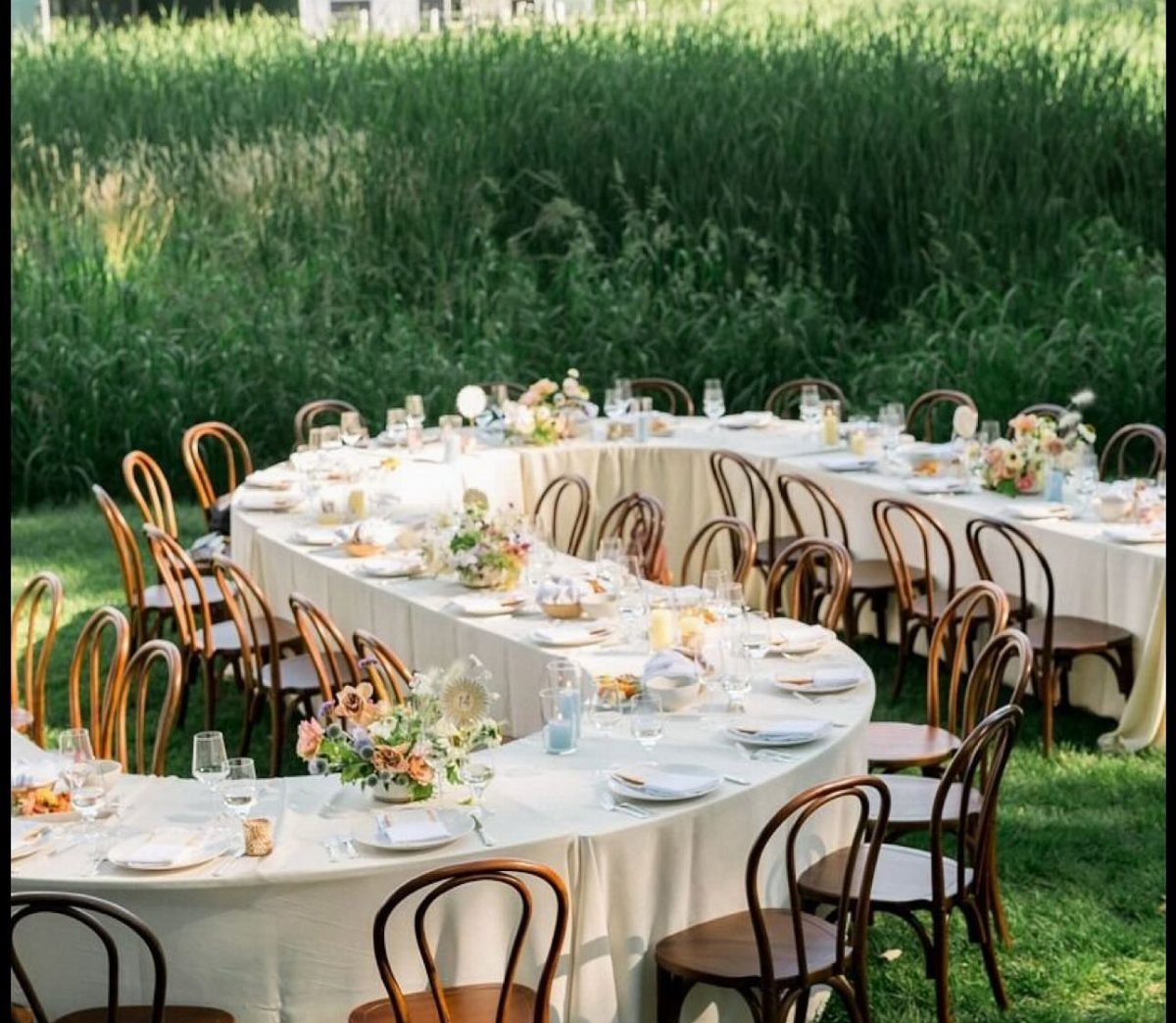 Curved white banquet table with floral arrangements and wooden chairs on green grass.