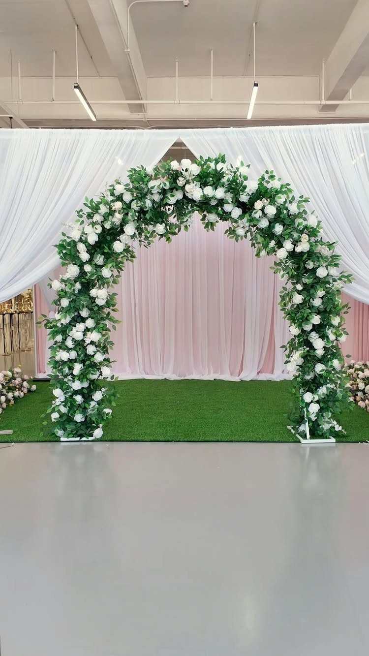 Floral arch with white roses and greenery against pink and white fabric drapes.