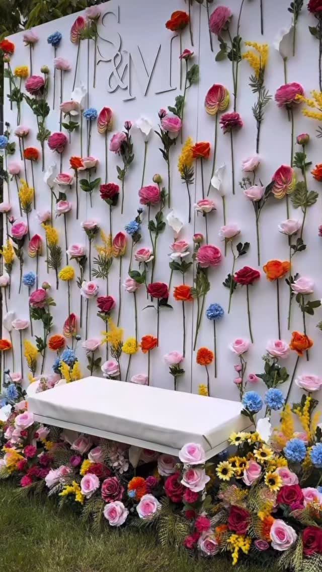 Colorful flower wall backdrop with a white bench and floral base on grass.