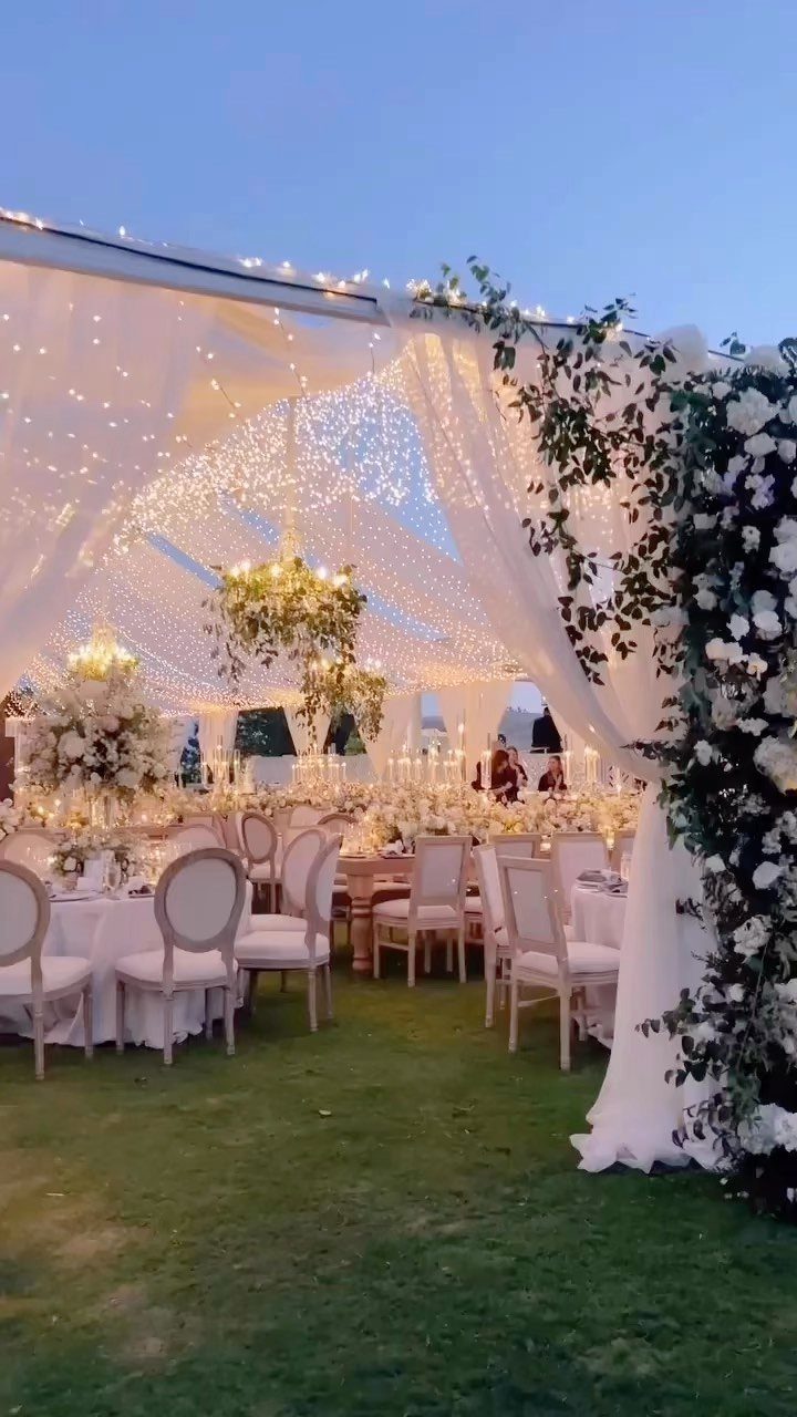 White wedding tent ceiling draped in fabric and glowing warm fairy lights over tables.