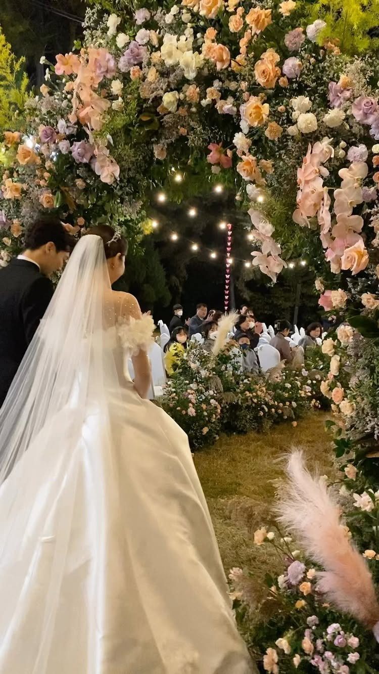 Bride and groom standing under a lush floral arch of pastel roses outdoors.