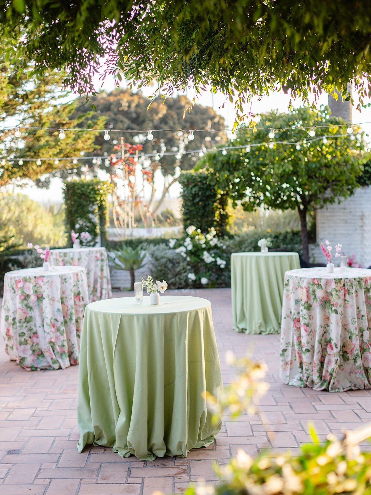 High-top cocktail tables with green and floral linens on a sunny brick patio.
