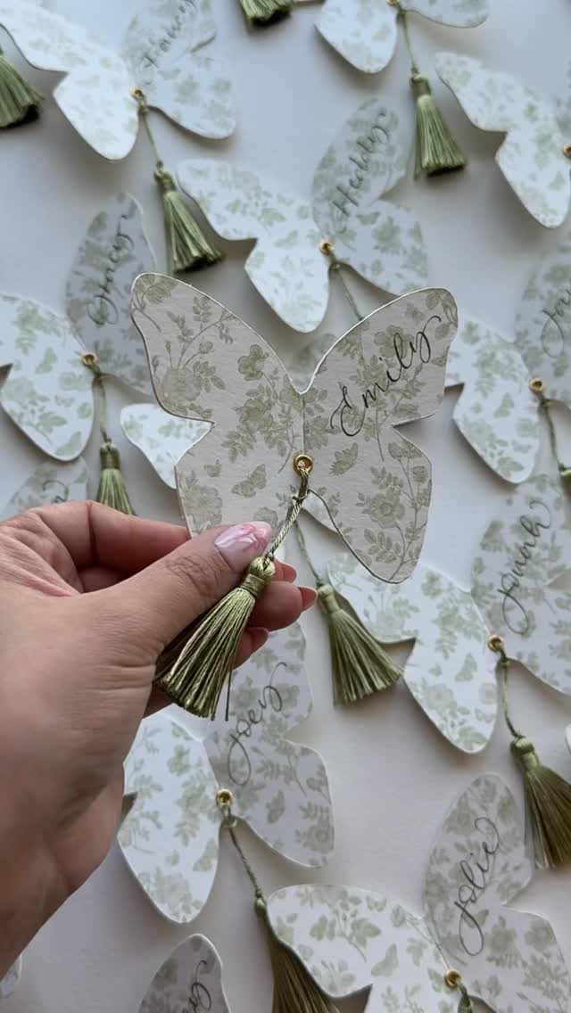 Butterfly-shaped place cards with green floral patterns and tassels for wedding table settings.
