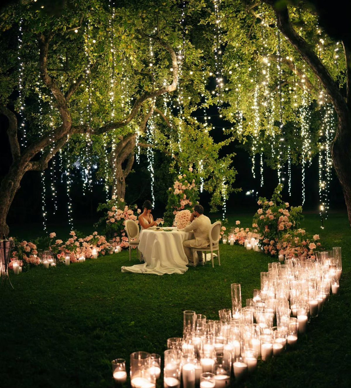 Couple dining under a tree with hanging fairy lights and candlelit grass path