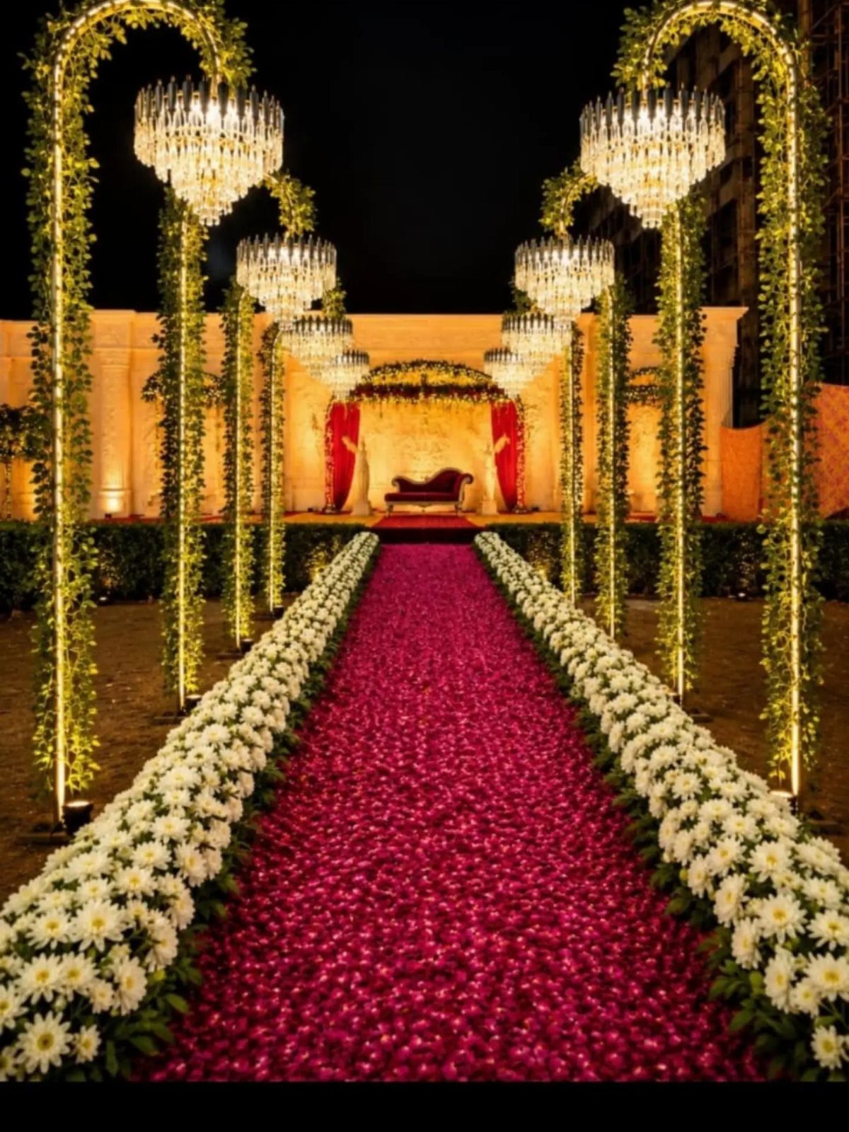 Red flower petal aisle bordered by white daisies under greenery arches and chandeliers.