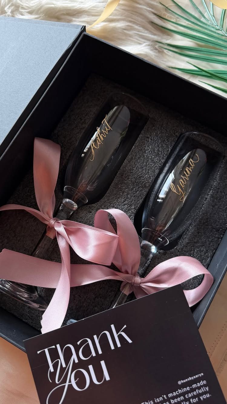 Two engraved champagne flutes in a black gift box with pink ribbons.
