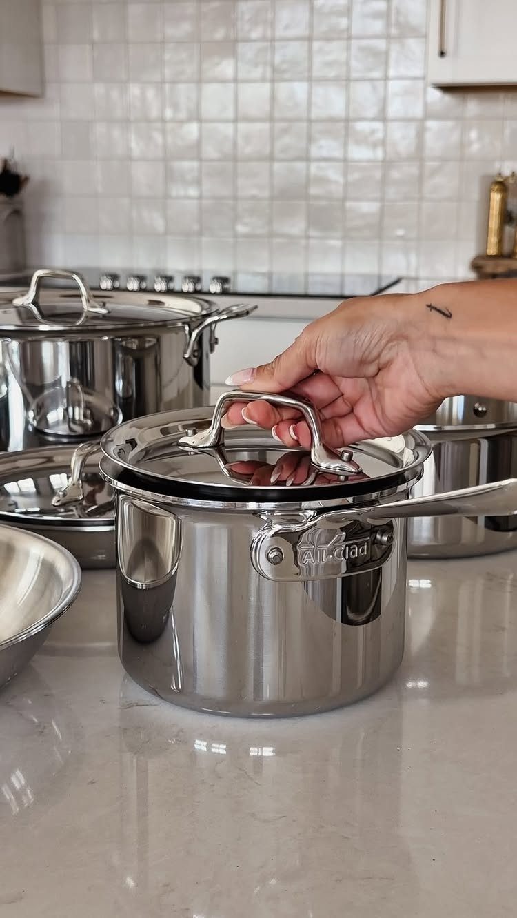 Stainless steel cookware set on a white marble kitchen counter with a hand.