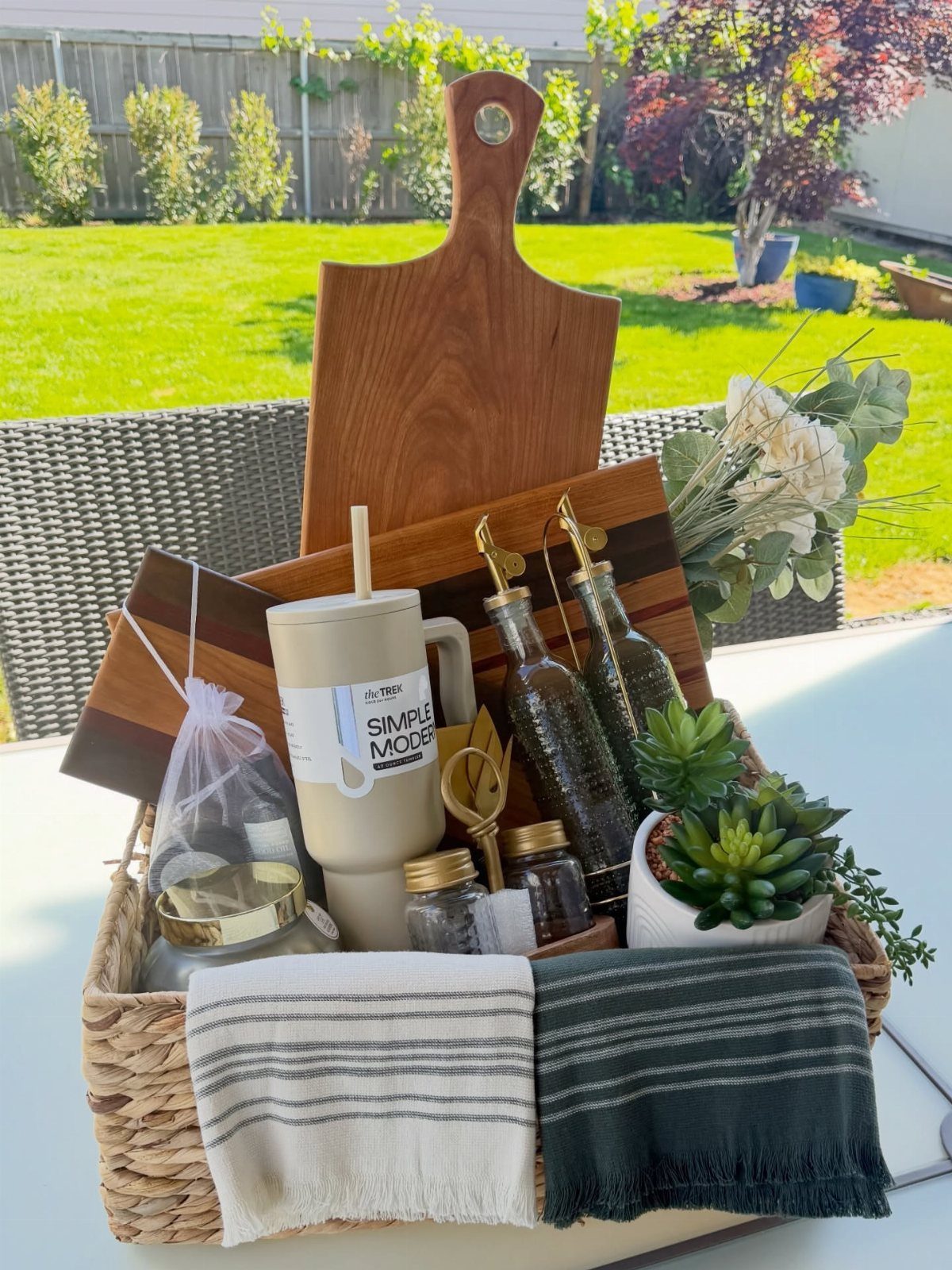 Wicker gift basket with wooden cutting board, mug, oil bottles, and towels outdoors.