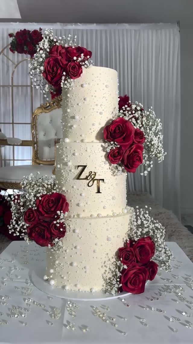 Three-tier white wedding cake with burgundy roses, baby's breath, and gold monogram initials.