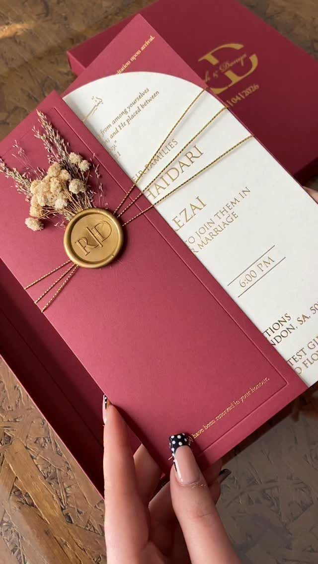 Burgundy wedding invitation with gold text, a gold wax seal, and dried flowers.