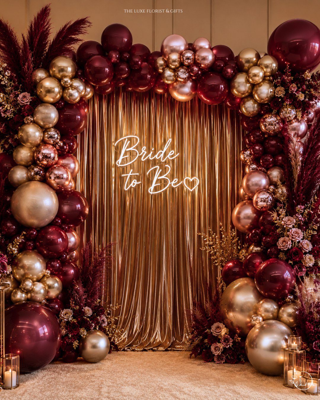 Gold shimmering curtain backdrop framed by burgundy and gold balloons and floral arrangements.