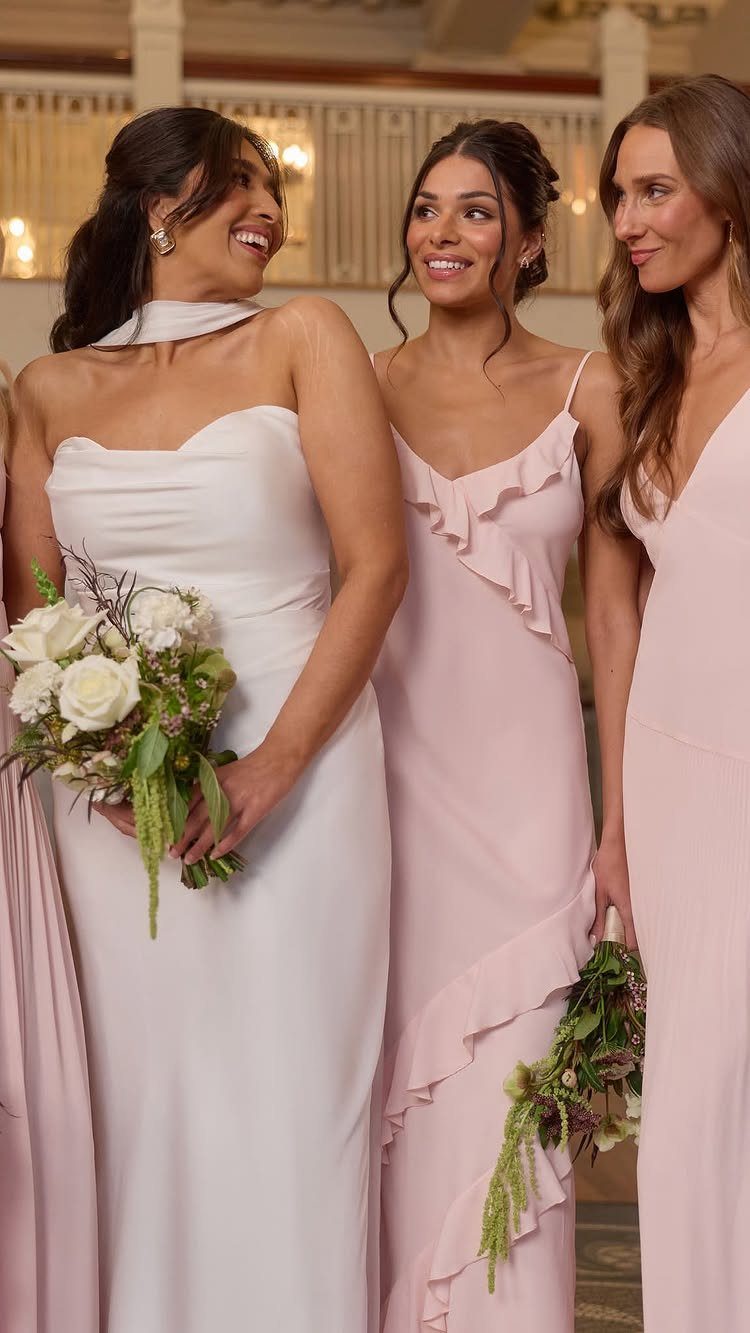 Three women in blush pink satin bridesmaid dresses holding white floral bouquets.