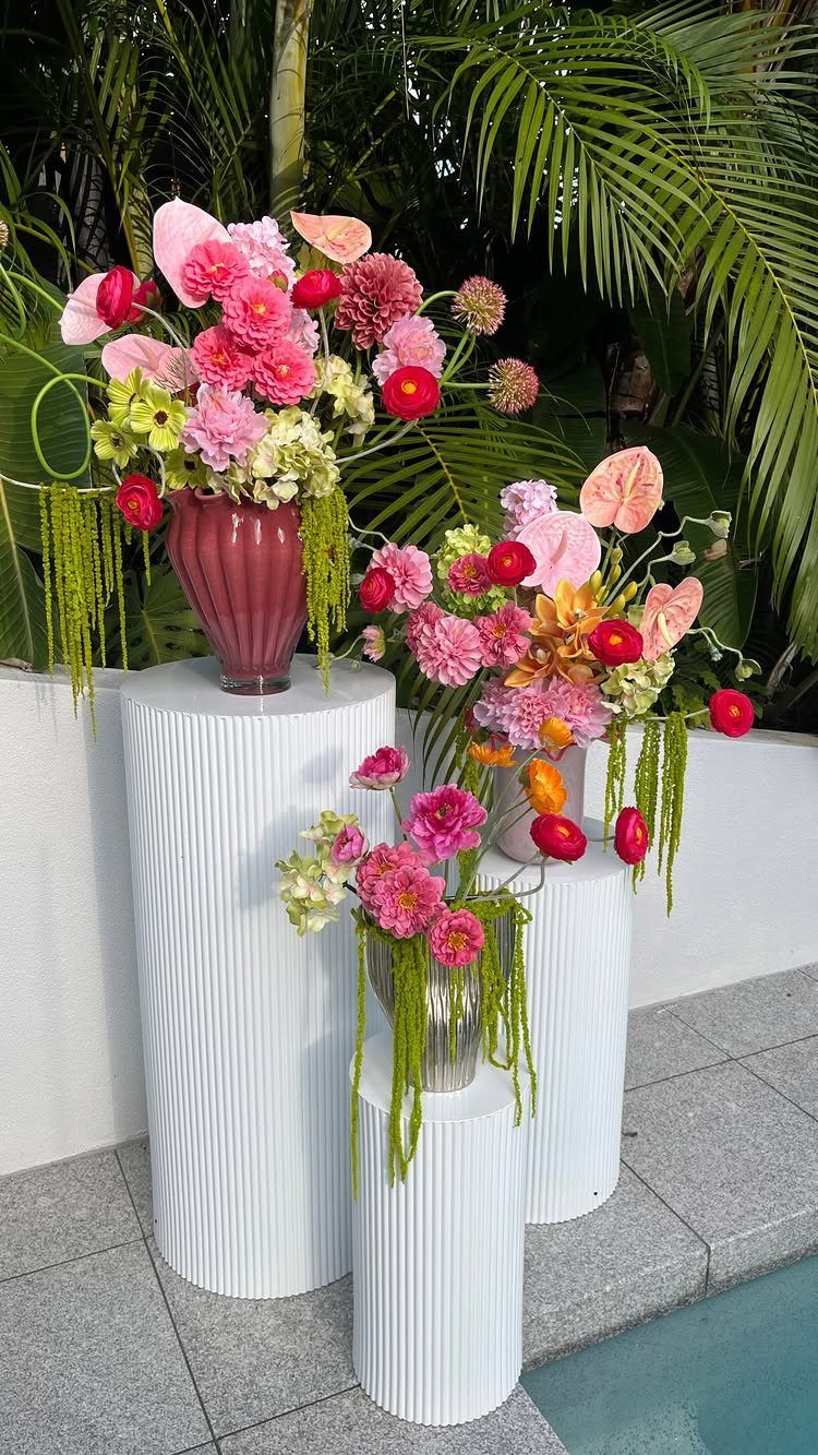 Pink and green floral arrangements in vases on white fluted pedestals outdoors.
