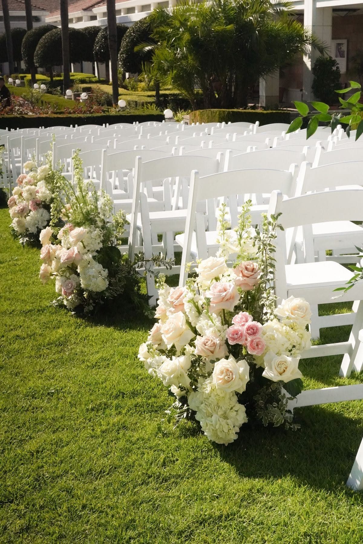 Pink and white floral arrangements lining a grass aisle between white wedding chairs.