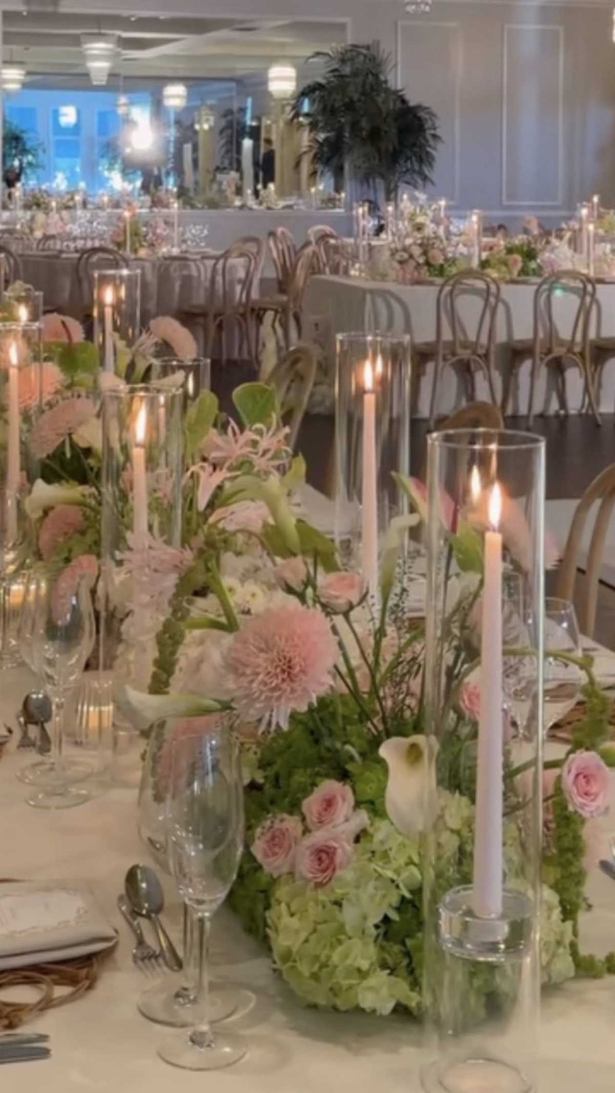 Pink and white taper candles in glass cylinders among pink and green floral centerpieces.