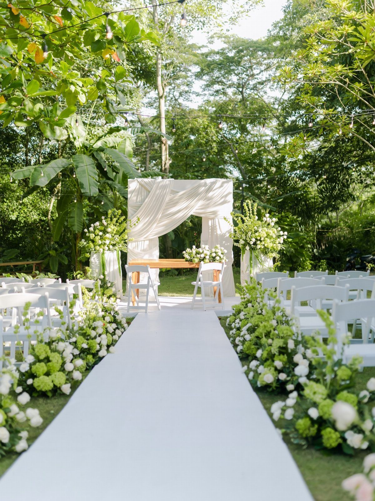Outdoor wedding ceremony setup with white aisle, floral borders, and draped altar.