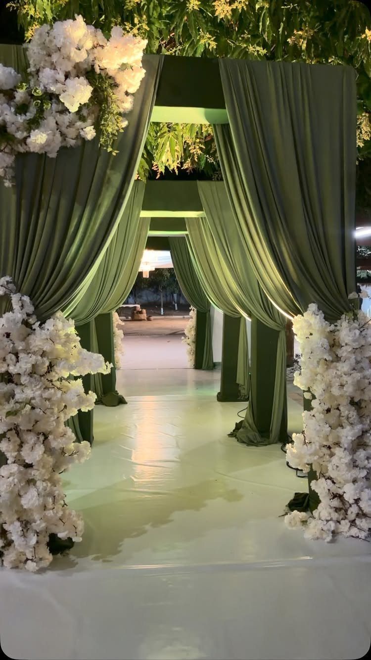 Olive green draped curtains and white flower arrangements lining a white wedding walkway.
