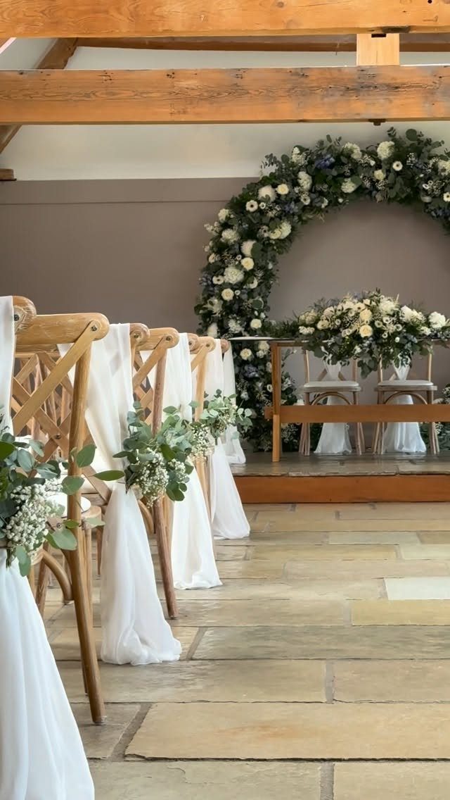 Wooden wedding chairs with white fabric and greenery facing a circular floral arch.