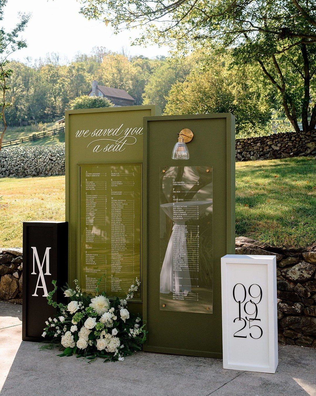 Olive green seating charts with gold calligraphy and white floral arrangements outdoors.