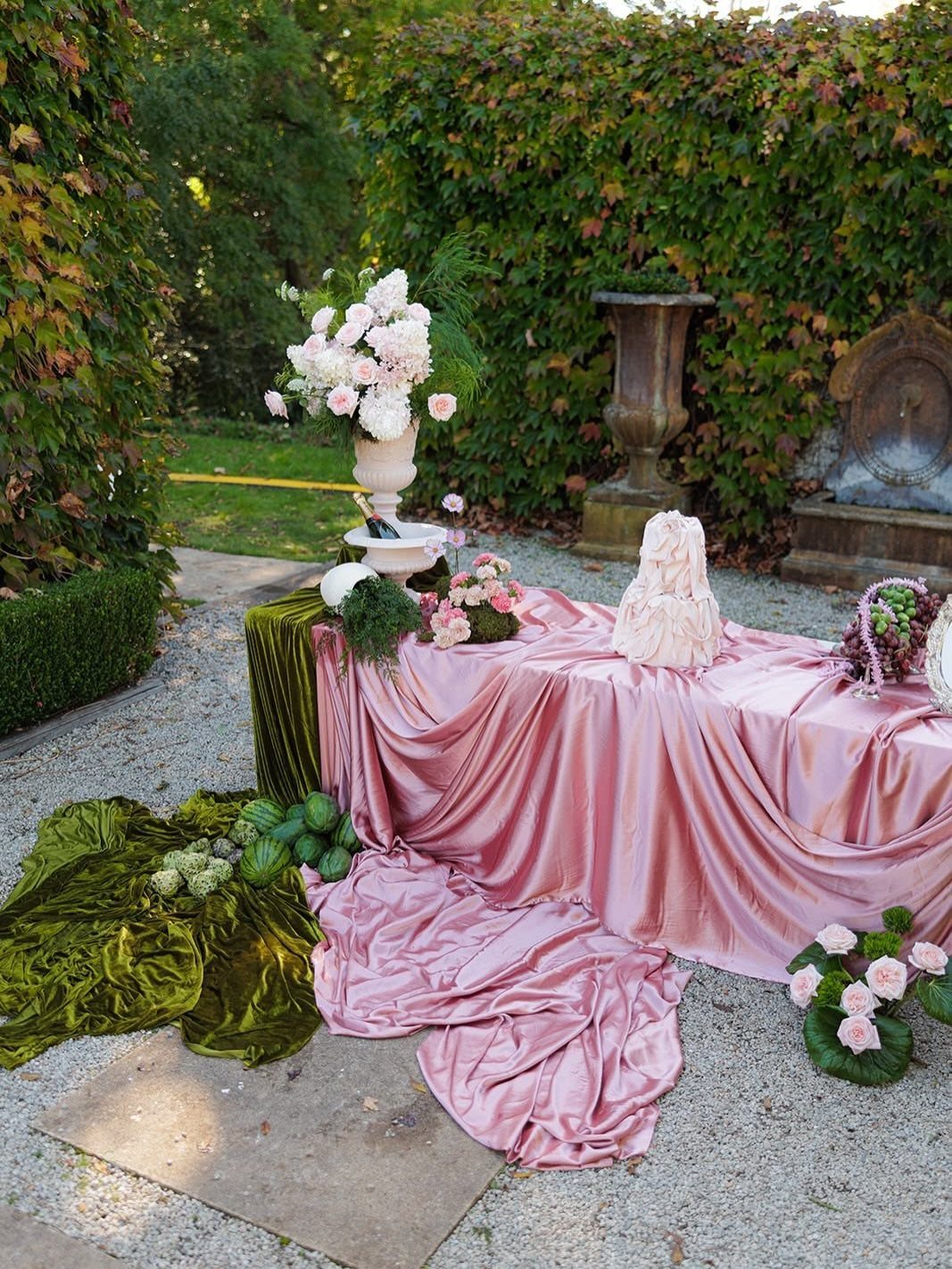 Pink satin draped table with white cake and green velvet accents in a garden