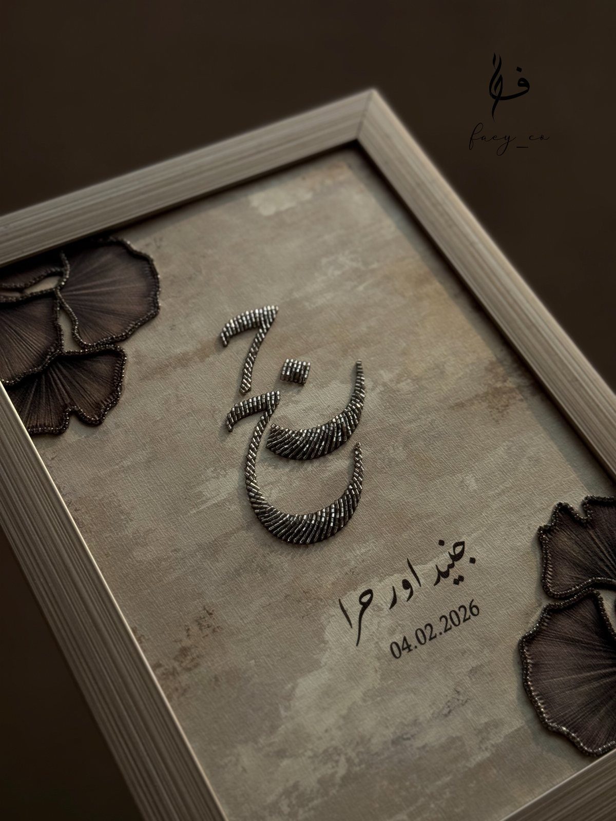 Framed embroidered Arabic calligraphy with floral accents and a date on beige fabric.