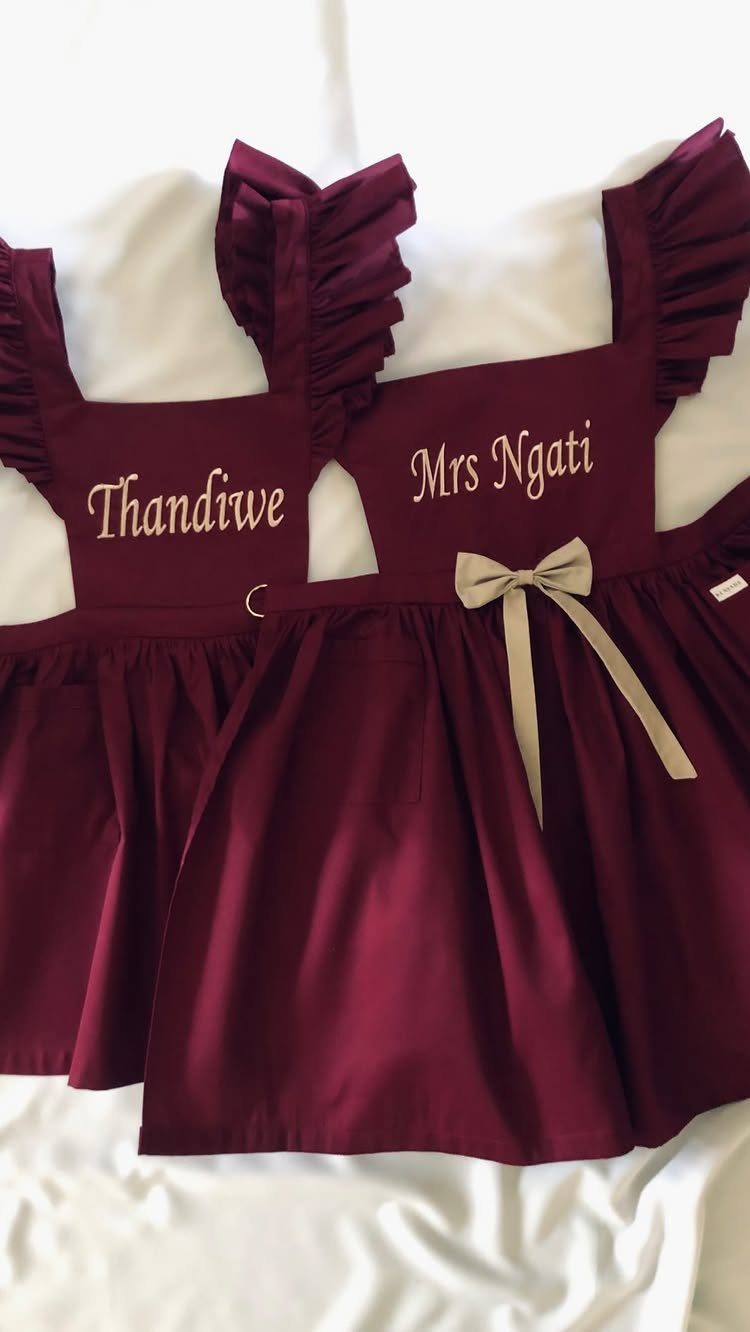 Two maroon personalized aprons with ruffled straps and gold embroidery on white fabric.