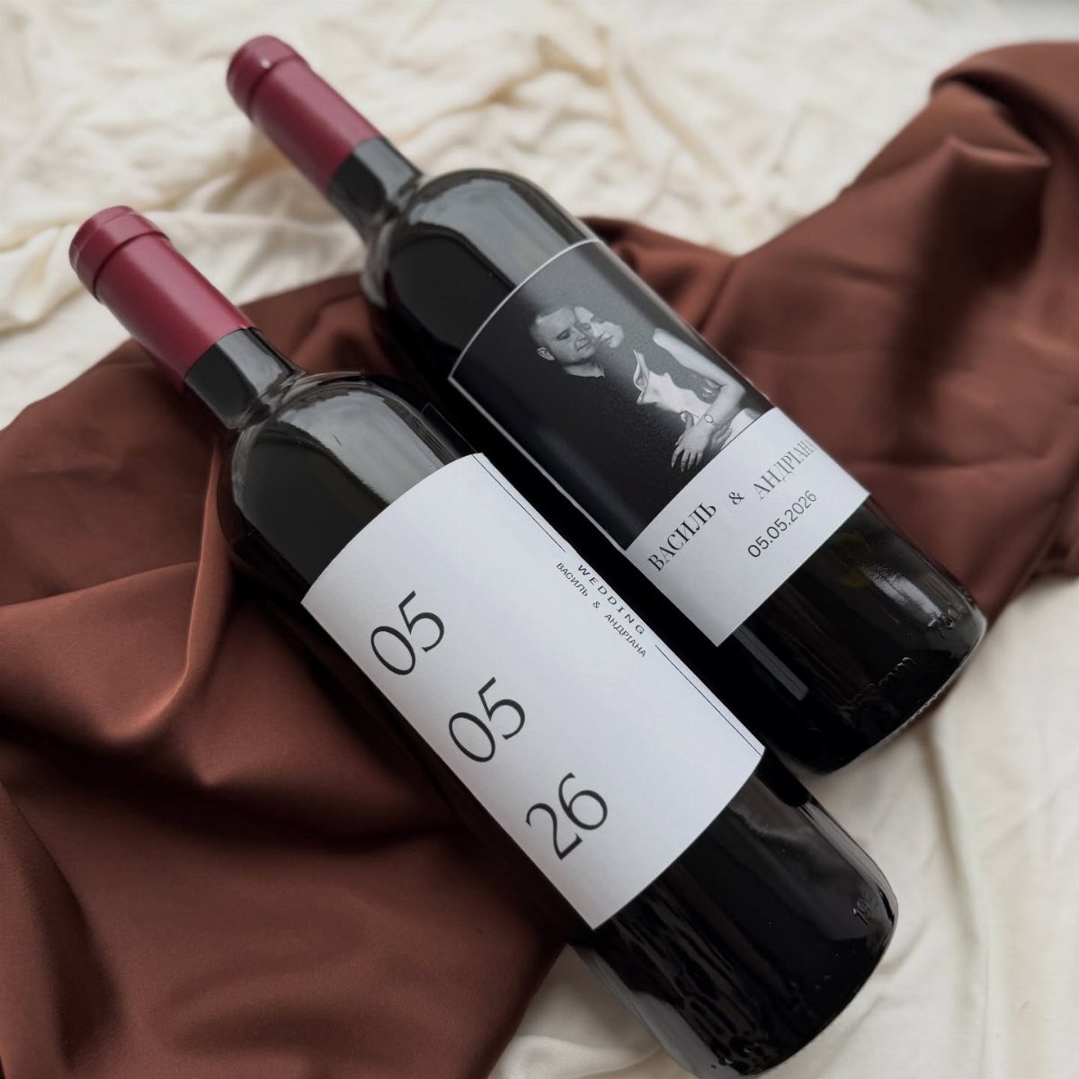 Two wine bottles with custom wedding labels resting on brown satin fabric.