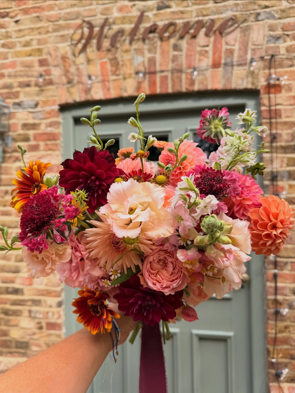 Wedding bouquet with burgundy, coral, and orange flowers against a brick wall background.