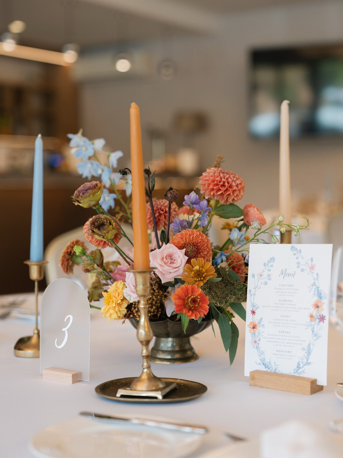 Floral centerpiece with orange and blue flowers and taper candles on wedding table.