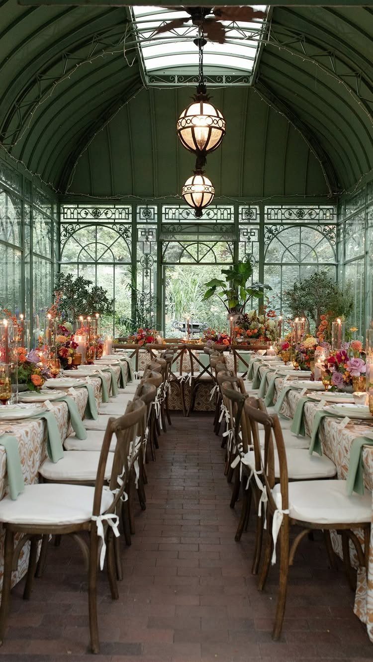 Long dining table with orange floral centerpieces inside a green glass conservatory.