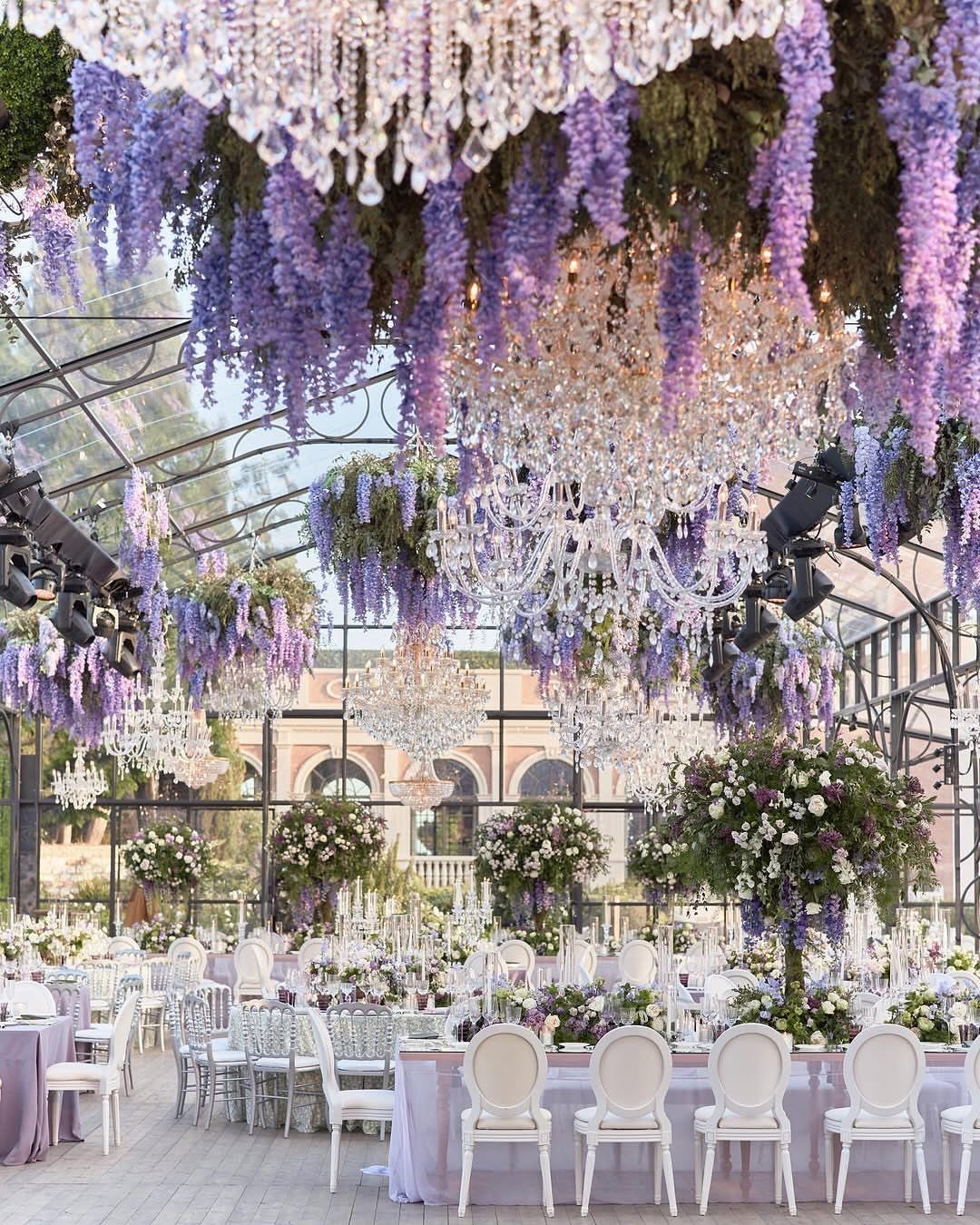 Outdoor wedding reception with hanging lilac wisteria, crystal chandeliers, and white chairs.