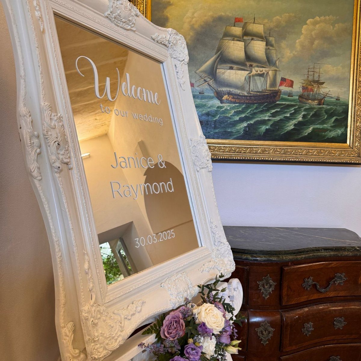 Ornate white mirror with wedding welcome text and lilac floral arrangement at base