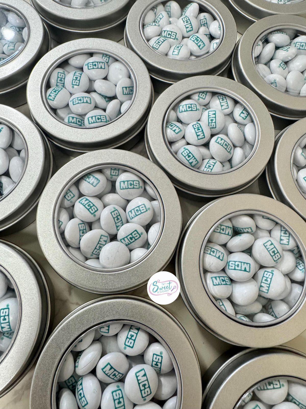 Silver metal tins filled with white M&M candies printed with teal letters