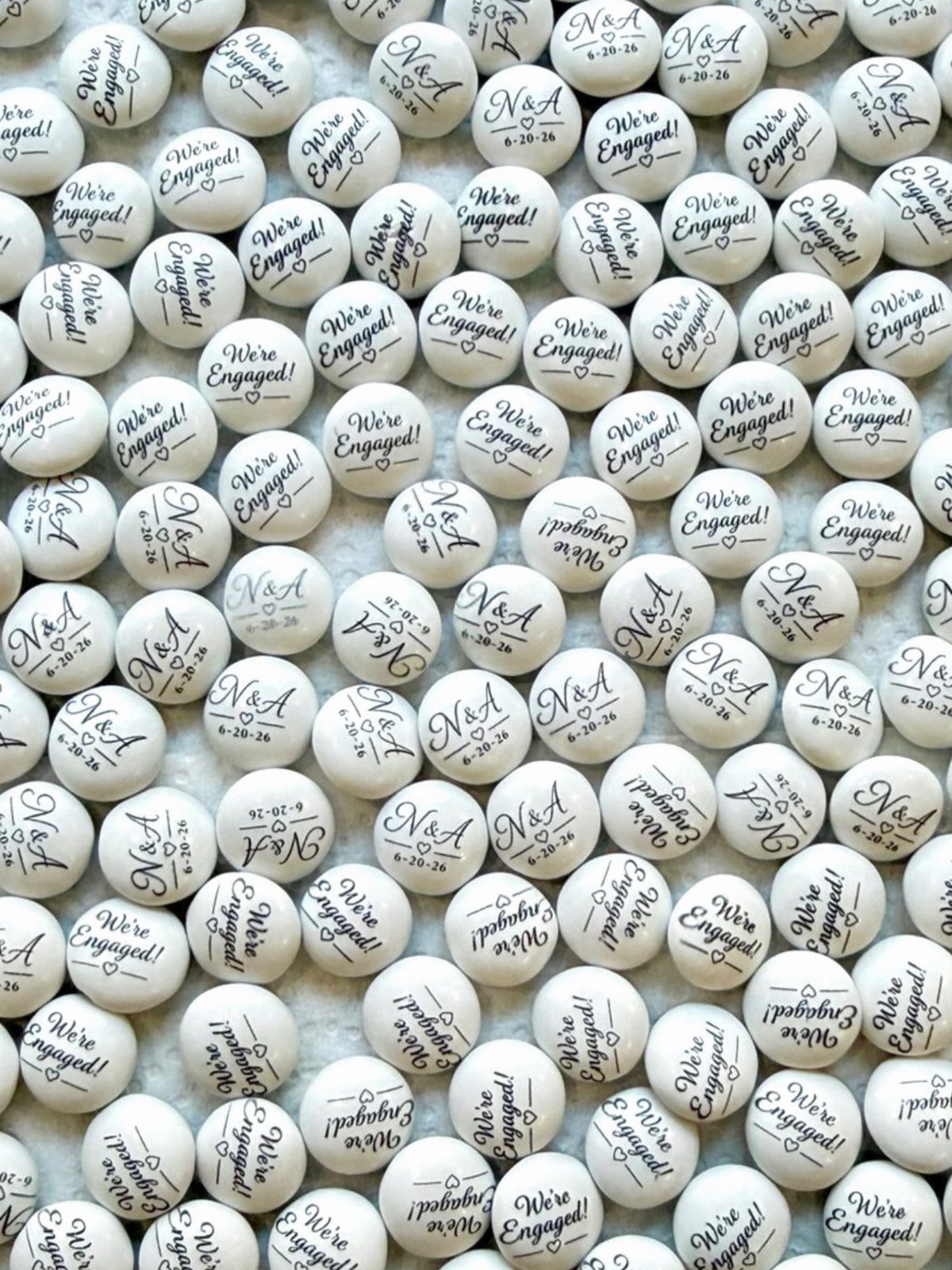 White M&M candies printed with engagement announcements and initials scattered on white.