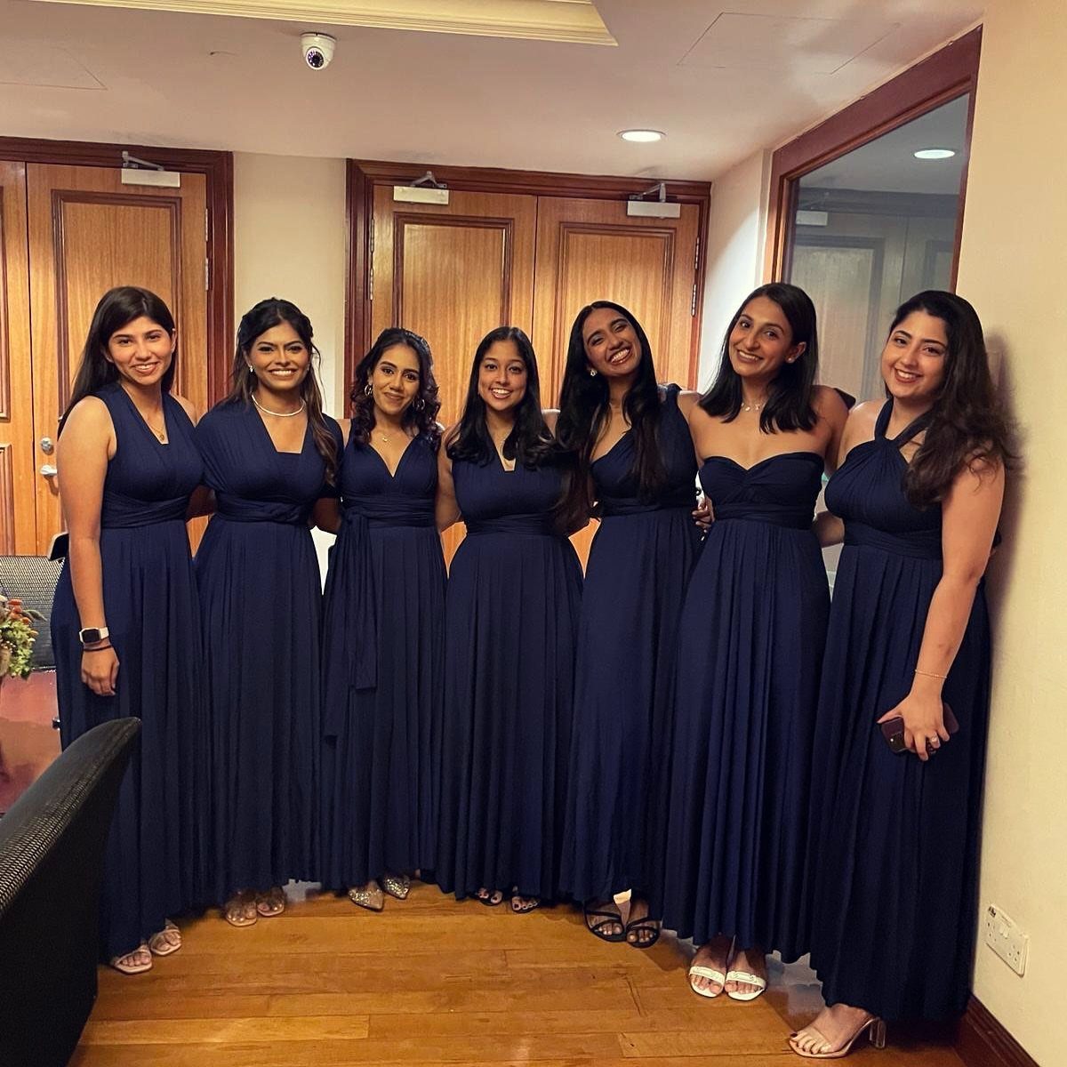 Seven women wearing long navy blue bridesmaid dresses posing together in a room.