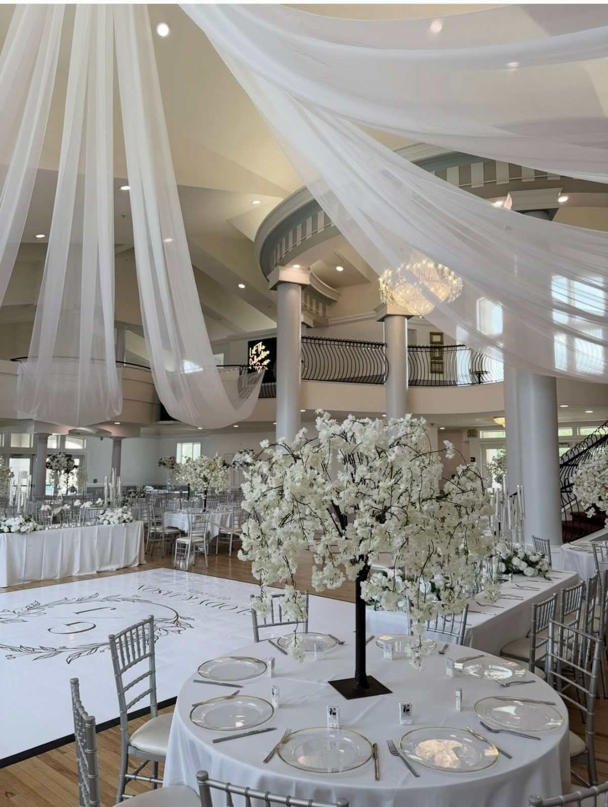 White wedding reception hall with draped ceilings, silver chairs, and white floral centerpieces.