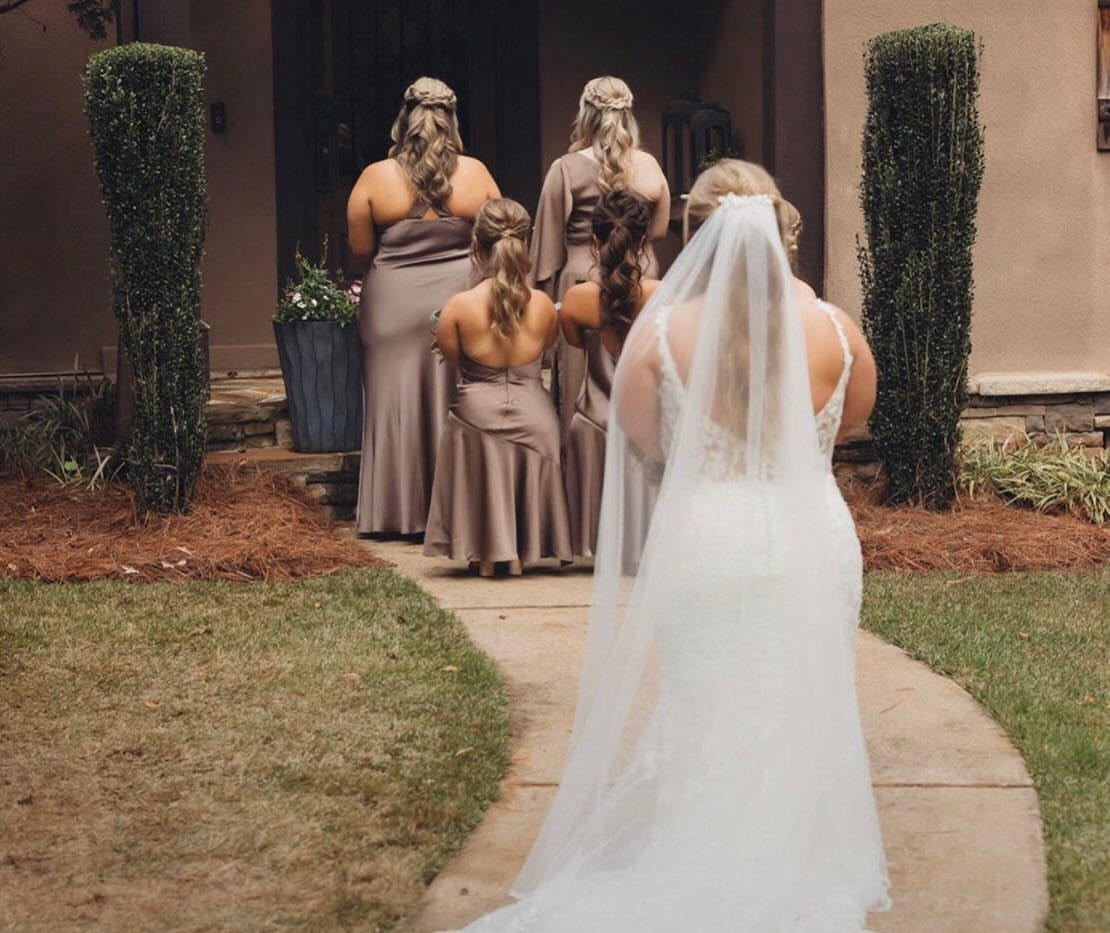 Bride in white lace gown and bridesmaids in taupe dresses walking toward a house.