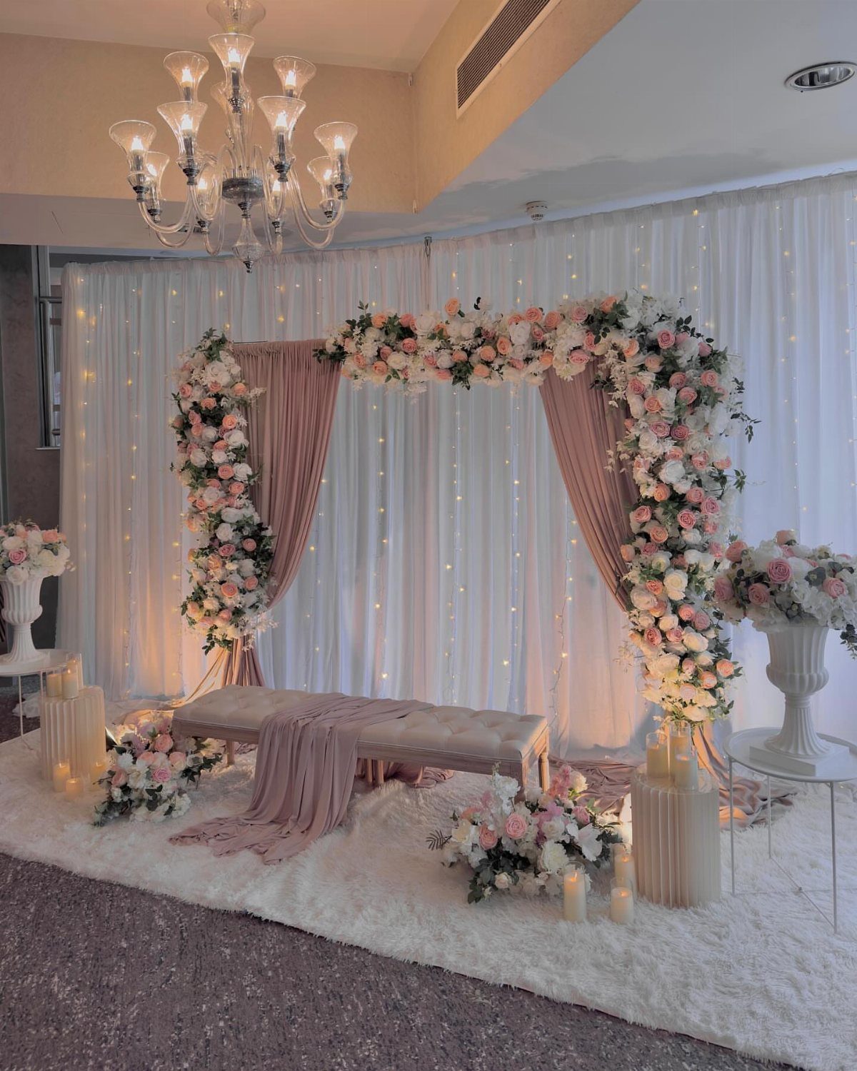Dusty rose wedding arch with pink flowers, white curtains, and a crystal chandelier.