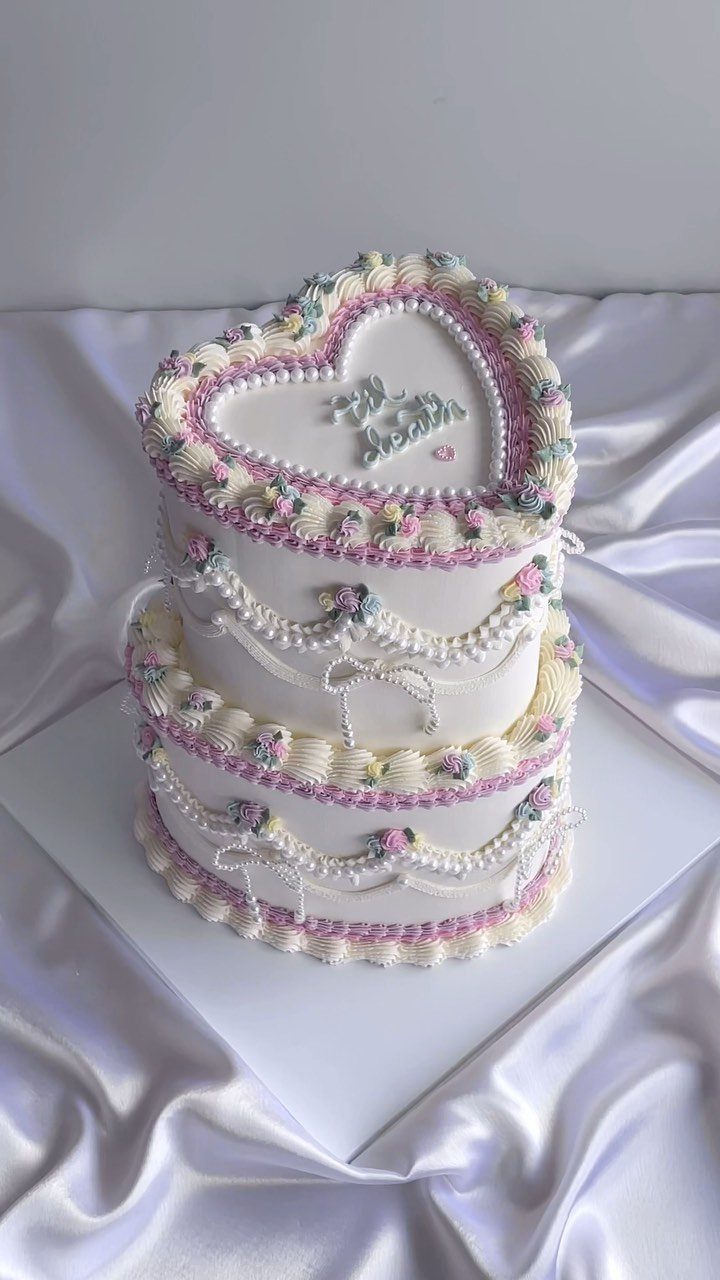 Heart-shaped two-tier white cake with pastel pink and purple frosting decorations on satin.
