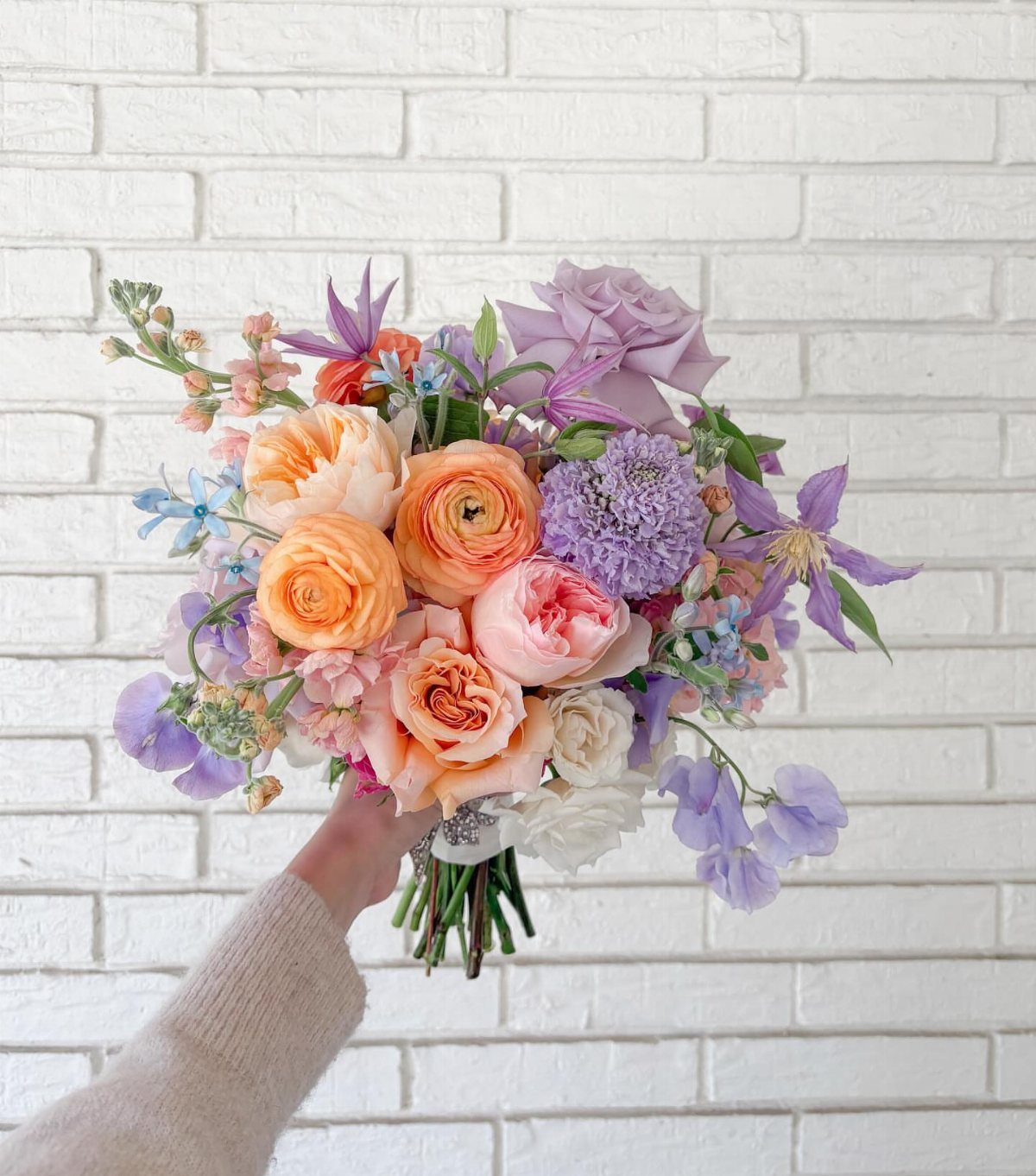 Bridal bouquet with peach and lavender flowers held against a white brick wall.