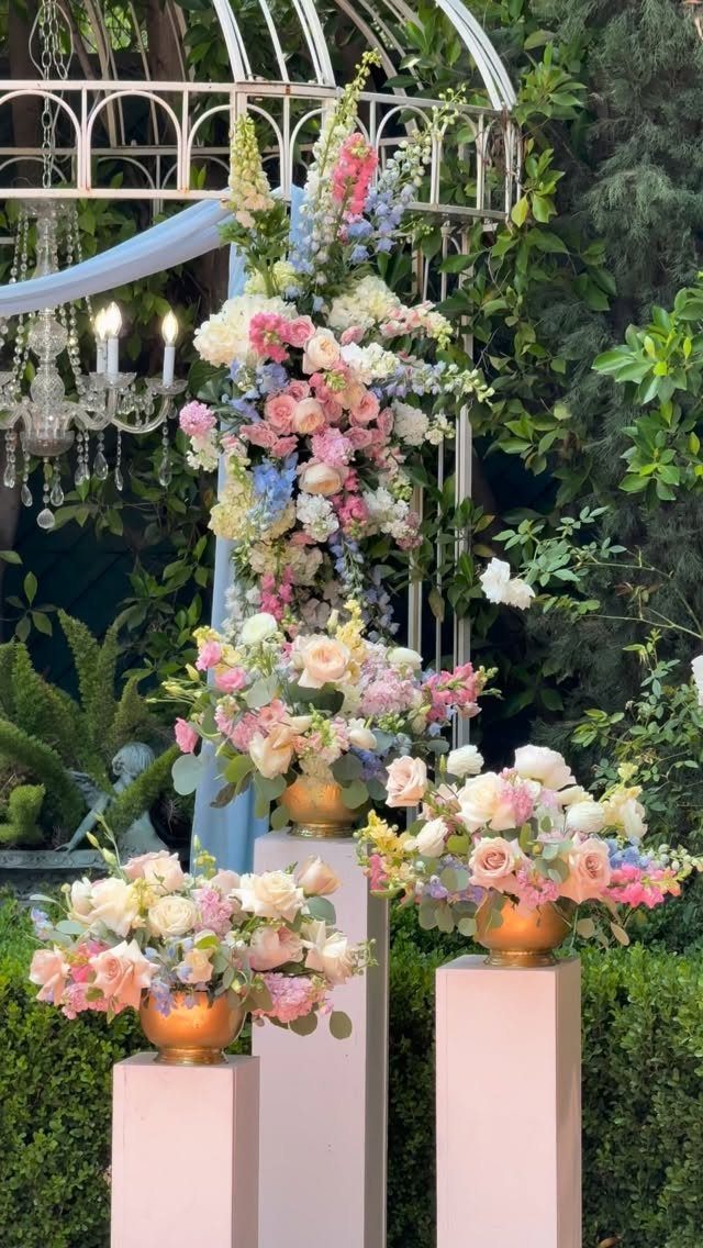 Pastel pink and blue floral arrangements on pink pedestals under a white arch.