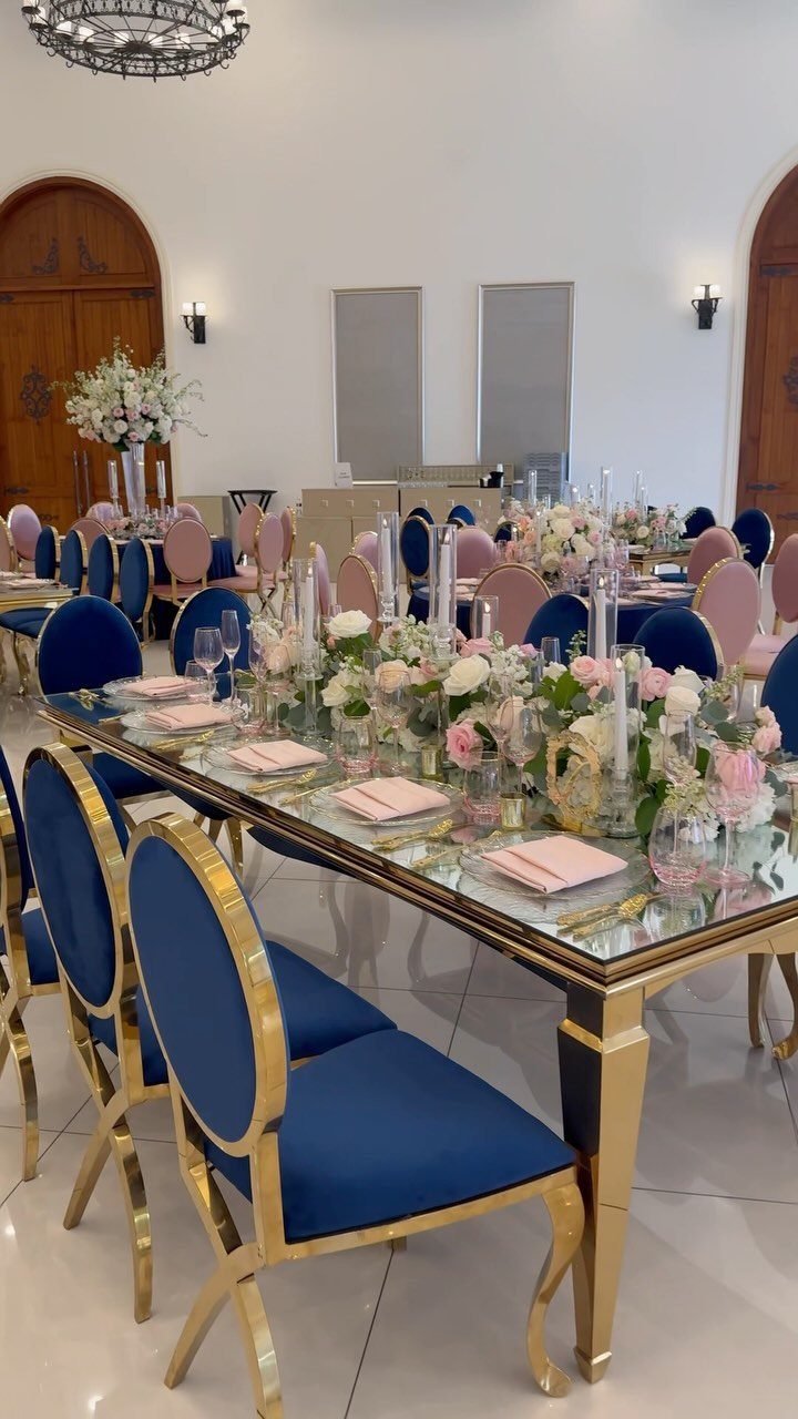 Glass dining table with pink floral centerpieces and navy blue velvet gold chairs.