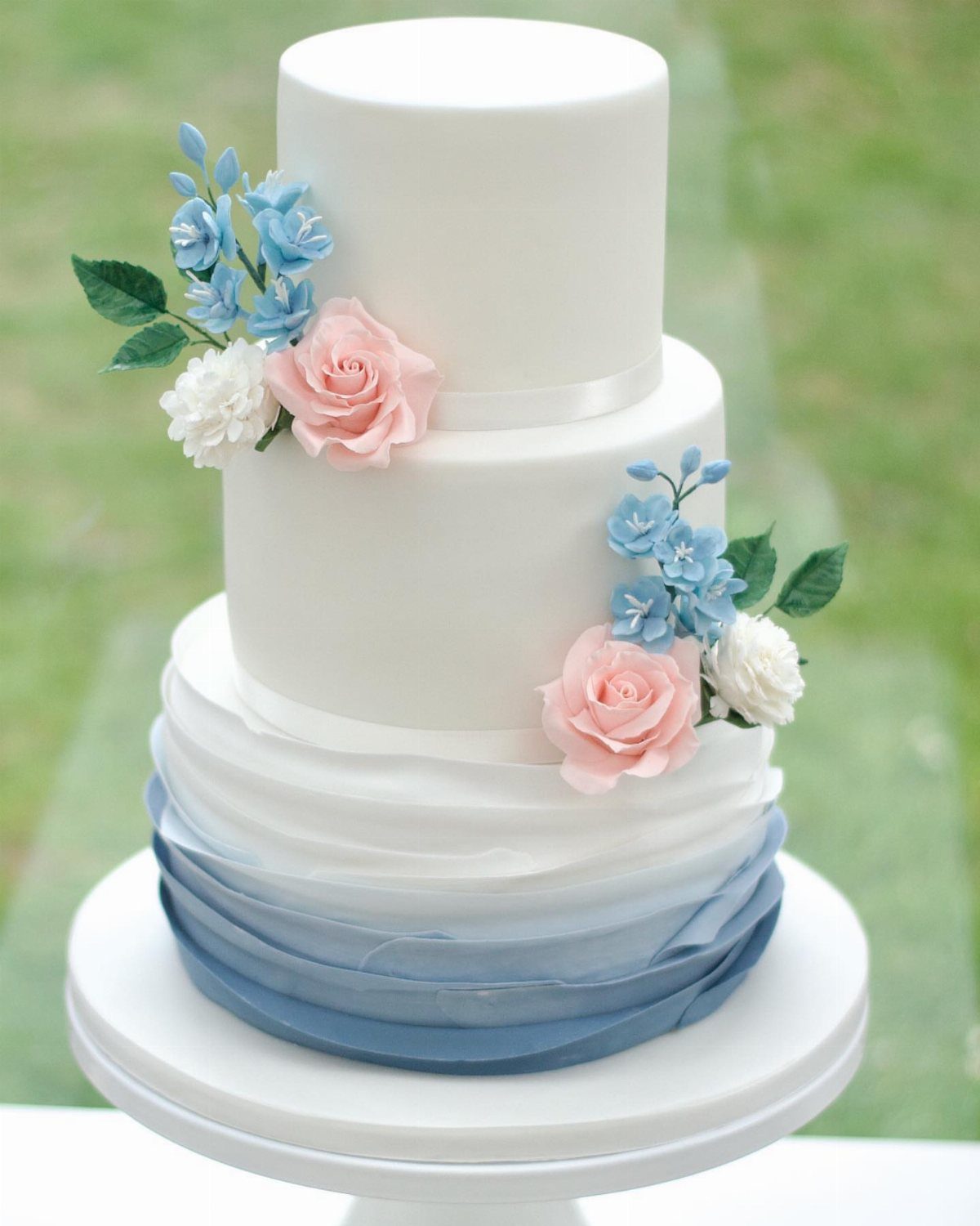Three-tier white wedding cake decorated with pink roses, blue flowers, and blue ruffles.