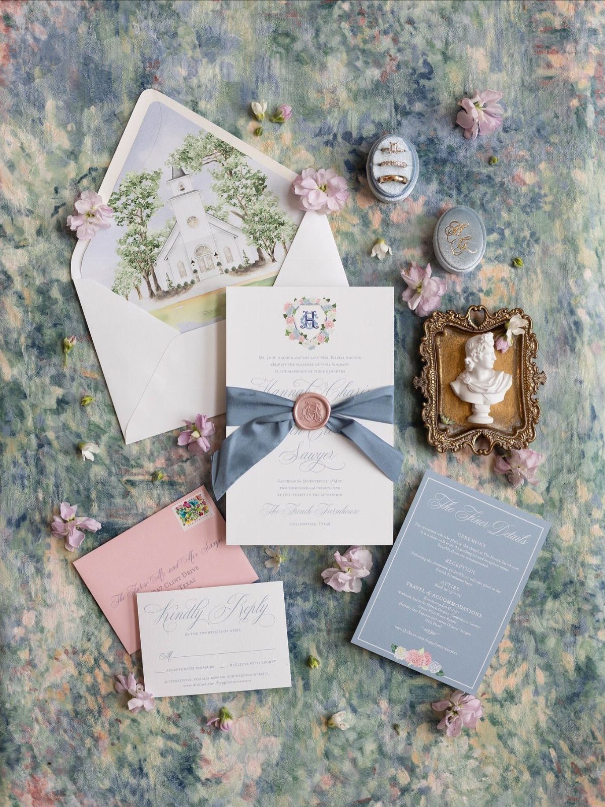 Wedding invitation suite with blue ribbon, pink envelope, and floral accents on textured background.