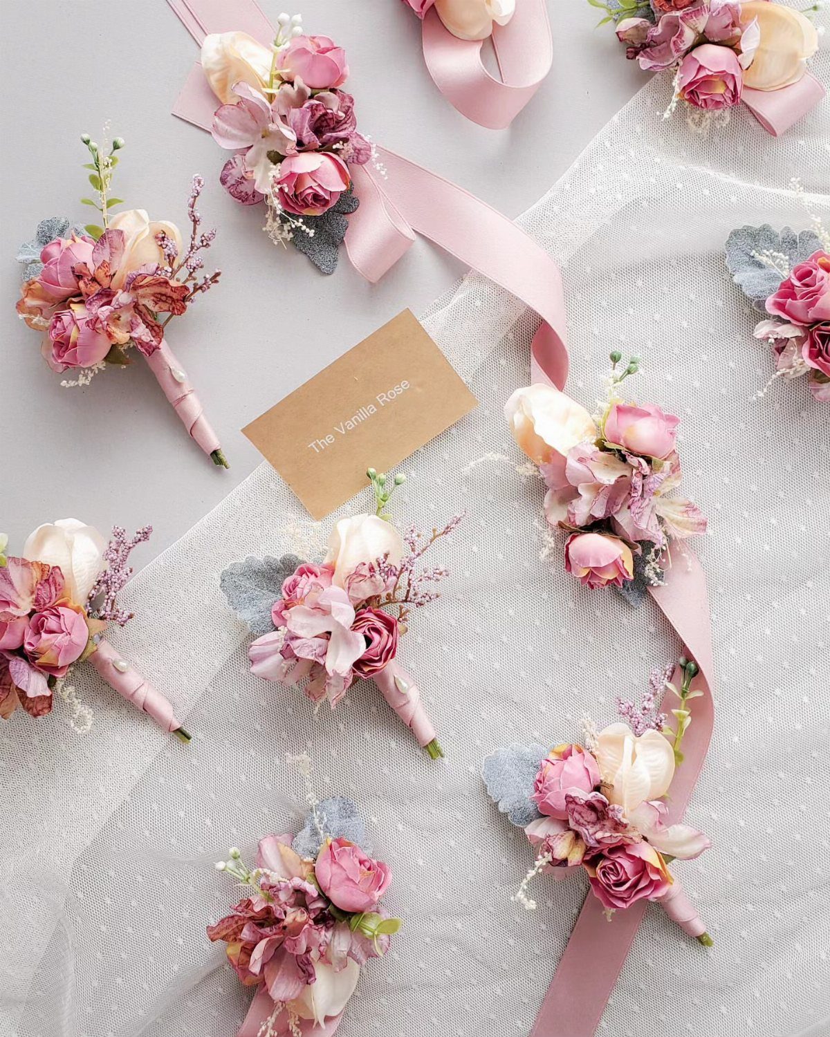 Dusty rose and cream floral boutonnieres arranged on white fabric with pink ribbons.