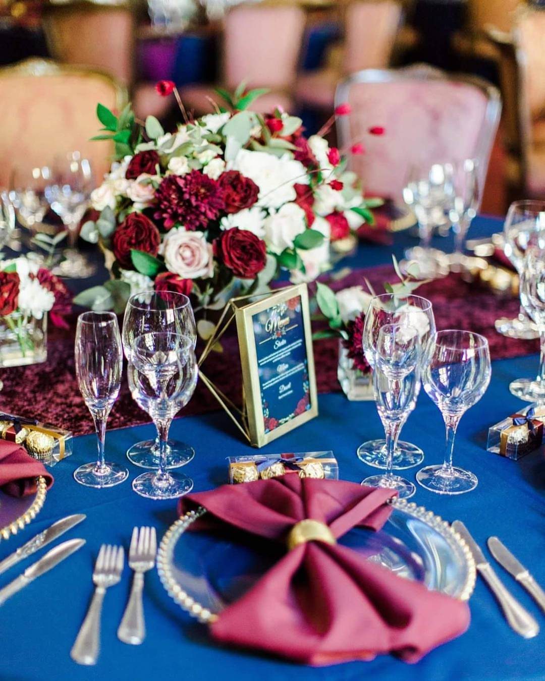 Navy blue wedding table with burgundy napkins, gold rings, and red floral centerpieces.