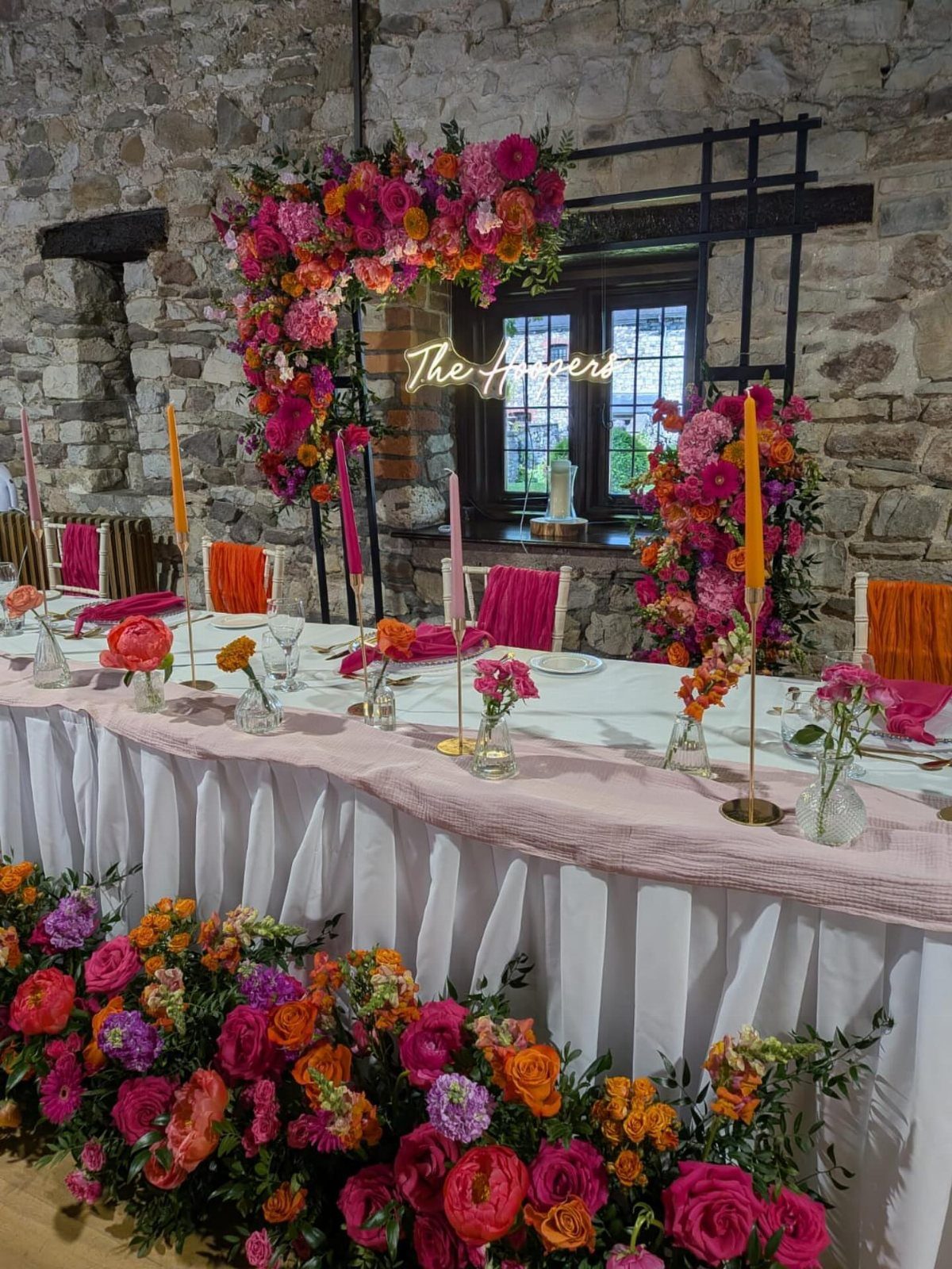 Wedding head table decorated with vibrant pink and orange flowers against stone walls.